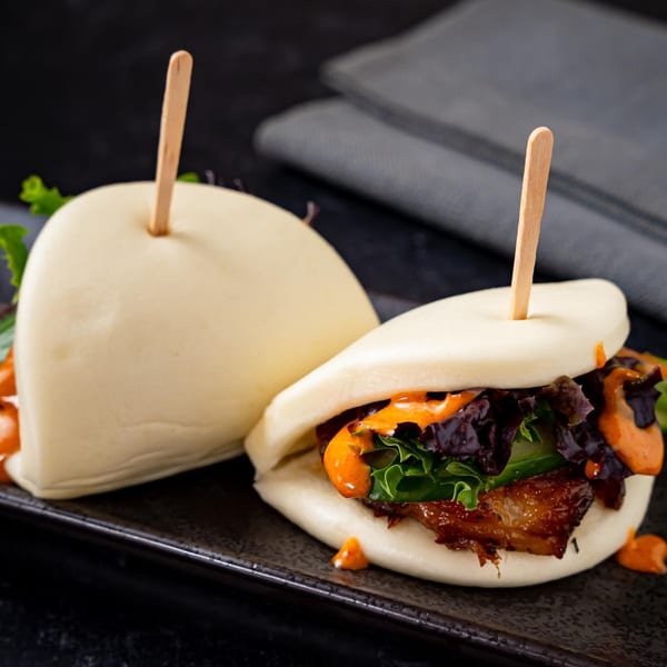 Two Shinjiru Ramen pork belly bao buns with lettuce and orange sauce on a dark plate.