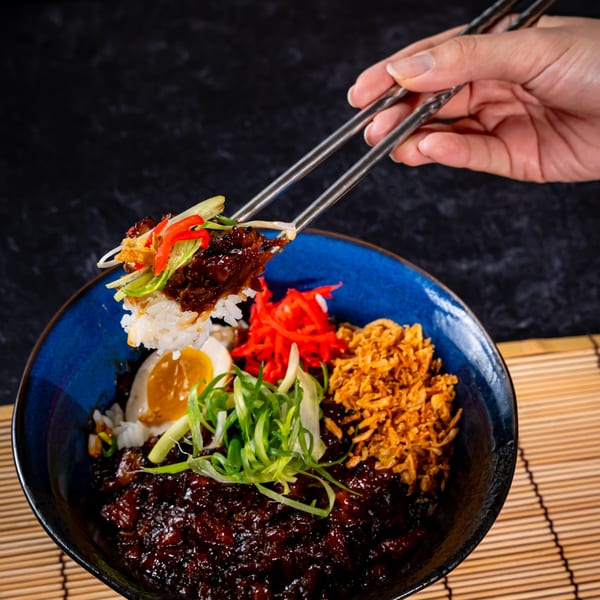 Chopsticks lifting rice and meat from a Shinjiru Ramen bowl with egg, scallions, and fried onions.