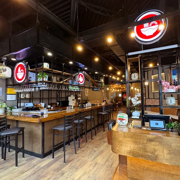 Interior of Shinjiru Ramen, featuring a long wooden bar with stools, ramen bowl logos, and decorative shelving.