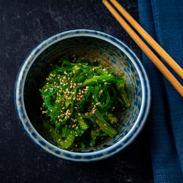 A blue bowl of green seaweed salad with sesame seeds, next to chopsticks and a blue napkin.
