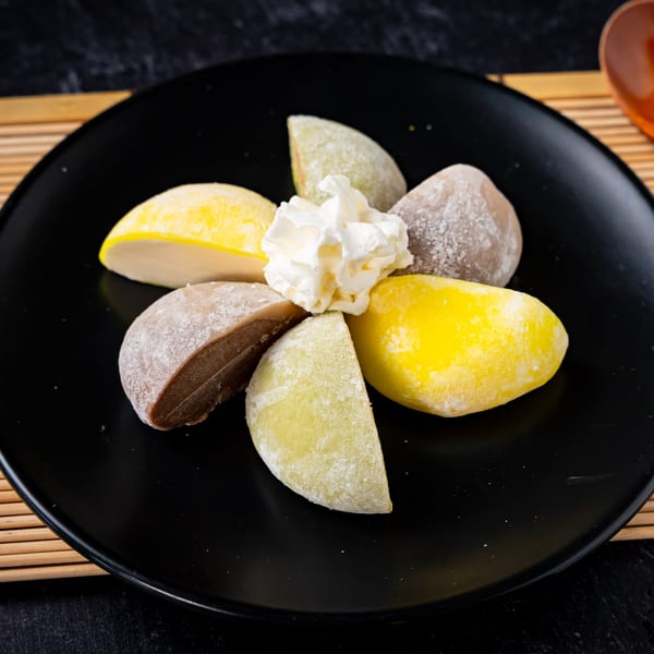 Assorted mochi ice cream, including yellow, green, and brown flavors, with whipped cream on a black plate.