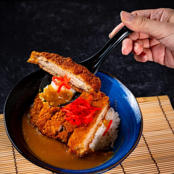 A hand lifts a piece of crispy tonkatsu from a bowl of curry rice with red ginger.