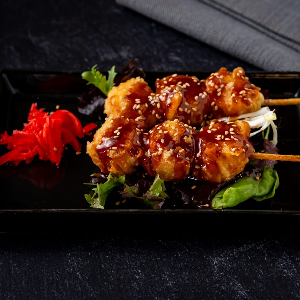 Shinjiru Ramen's fried chicken skewers with teriyaki sauce, sesame seeds, and pickled ginger on a black plate.