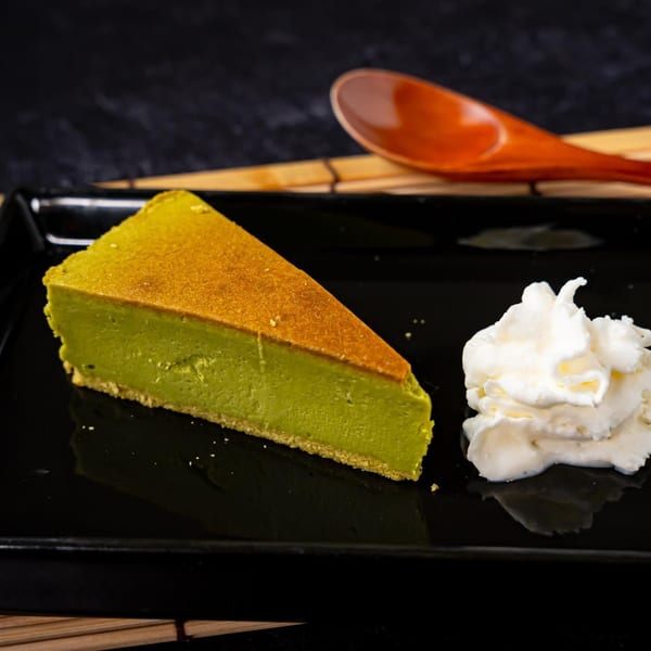 Slice of green matcha cheesecake with whipped cream on a black plate, with a wooden spoon.