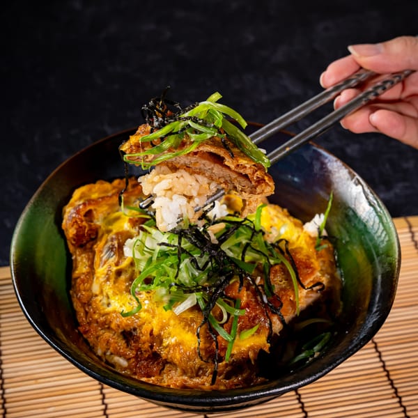 Chopsticks lifting a piece of crispy tonkatsu and rice from a bowl of Katsudon.