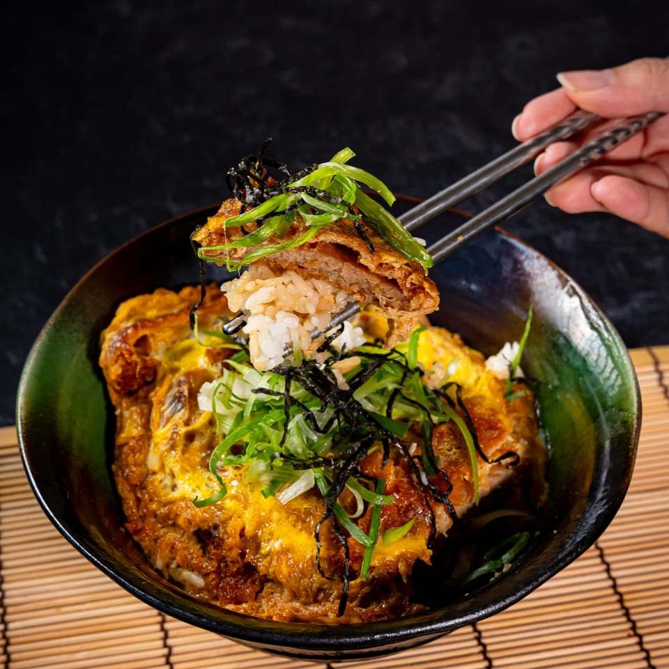 Chopsticks lifting a piece of crispy tonkatsu and rice from a bowl of Katsudon.