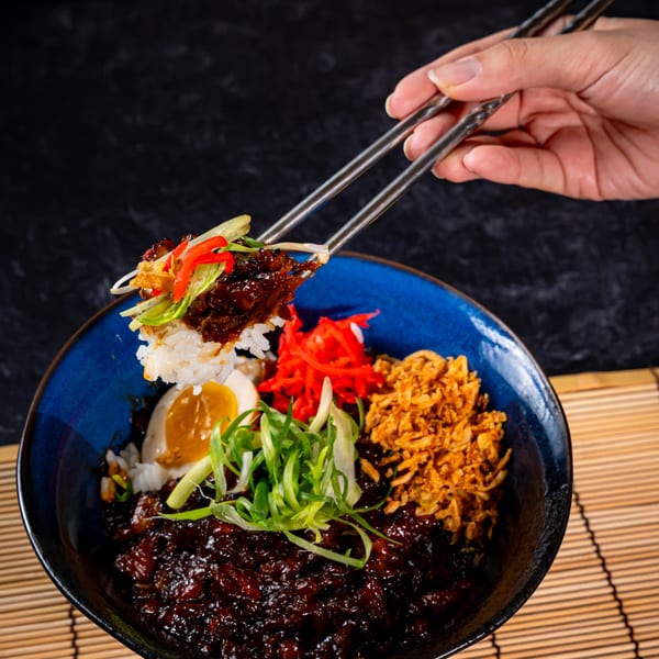Chopsticks lifting rice and meat from a Shinjiru Ramen bowl with egg, scallions, and fried onions.