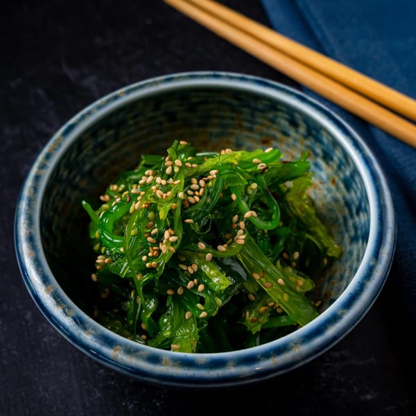 Shinjiru Ramen's vibrant green seaweed salad with sesame seeds in a blue bowl, with chopsticks.