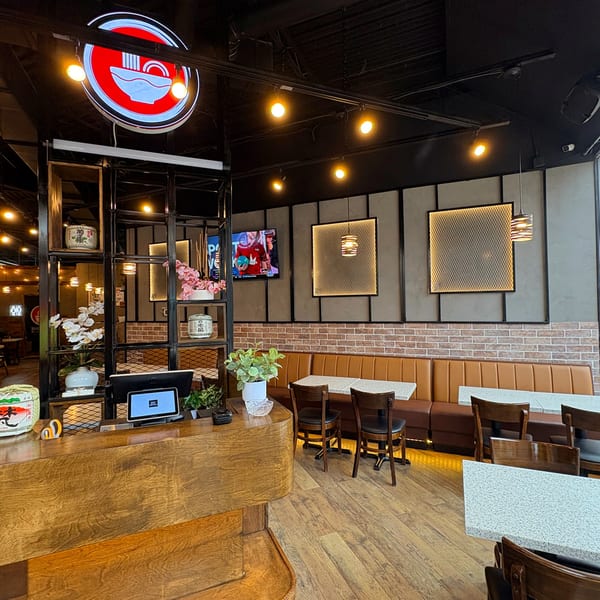 Interior of Shinjiru Ramen with a wooden counter, booths, tables, and a neon ramen bowl sign.