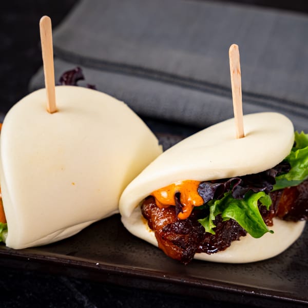 Two Shinjiru Ramen bao buns with pork belly, lettuce, and orange sauce on a dark plate.