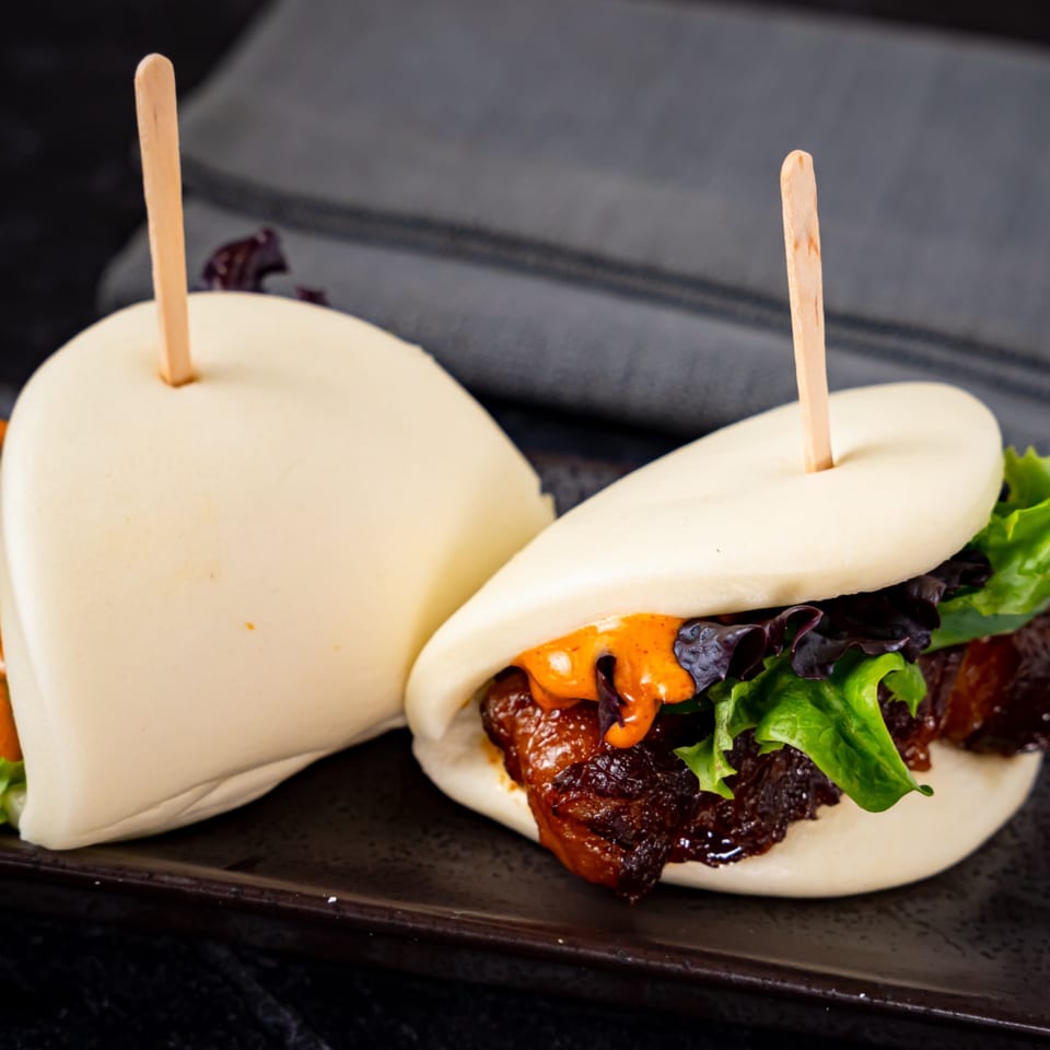 Two Shinjiru Ramen bao buns with pork belly, lettuce, and orange sauce on a dark plate.