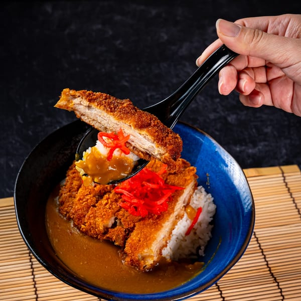 A hand lifts a piece of crispy tonkatsu and curry from a blue bowl of rice.