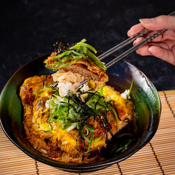 Chopsticks lifting a piece of tonkatsu and rice from a bowl of Katsudon, garnished with green onions.