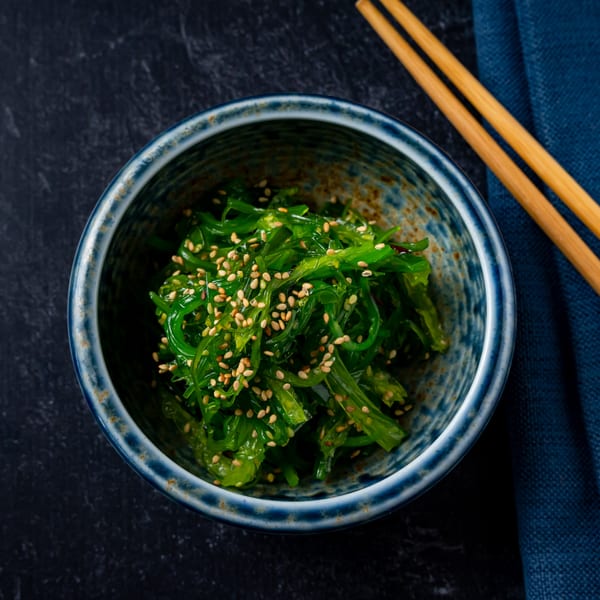 A blue ceramic bowl filled with green seaweed salad topped with sesame seeds, next to chopsticks.