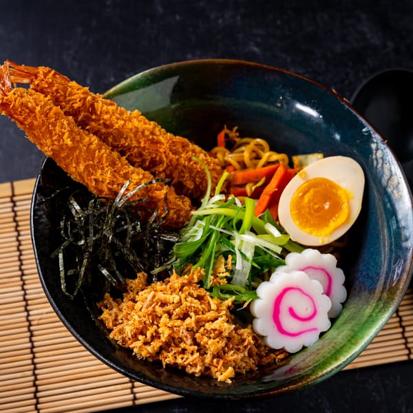 Shinjiru Ramen bowl with crispy shrimp, egg, narutomaki, seaweed, and green onions on a bamboo mat.