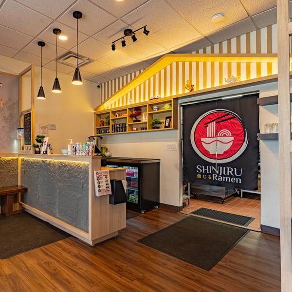 Shinjiru Ramen restaurant interior with a stone-front counter, wooden shelves, and a branded curtain.