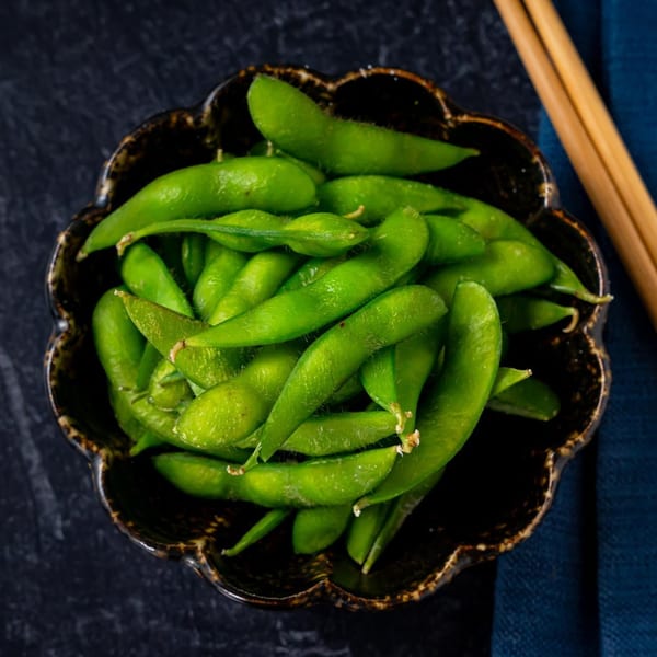 A dark, ornate bowl filled with vibrant green edamame pods, with chopsticks and a blue napkin nearby.