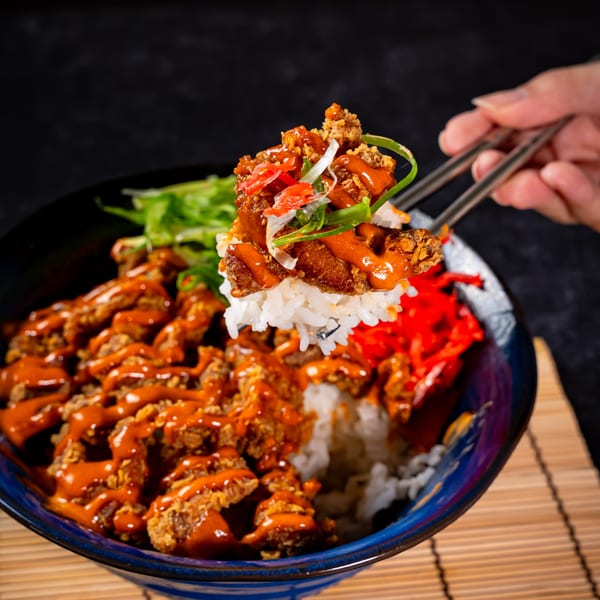Chopsticks lift a piece of crispy chicken with sauce and rice from a colorful bowl.